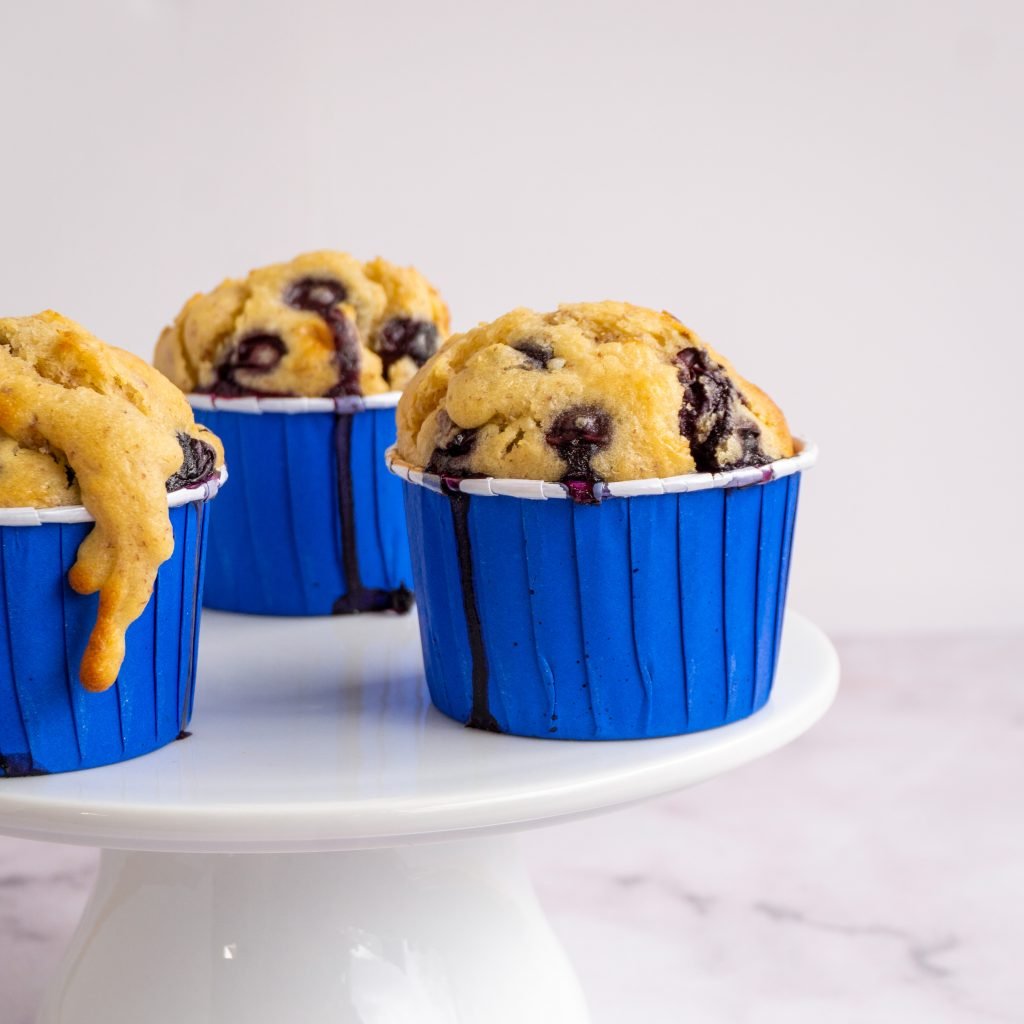 Dieses Bild zeigt drei Blaubeer Muffins auf einer Tortenplatte von vorne
